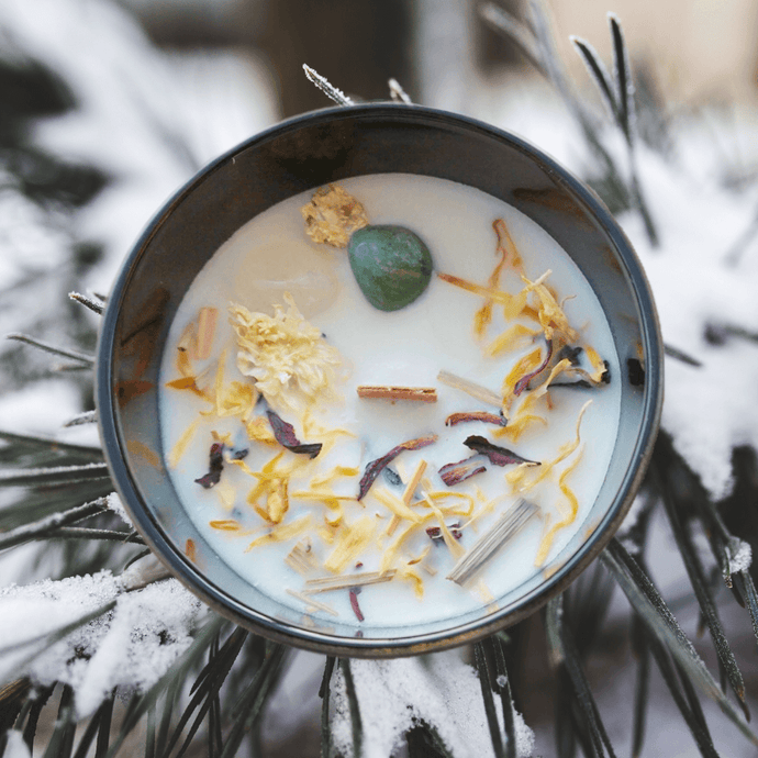 A picture of a large white candle sprinkled with Lemongrass, ginger root, rosella hibiscus flower & calendula flower and two crystals Green Aventurine and clear Quartz cimbedded in it